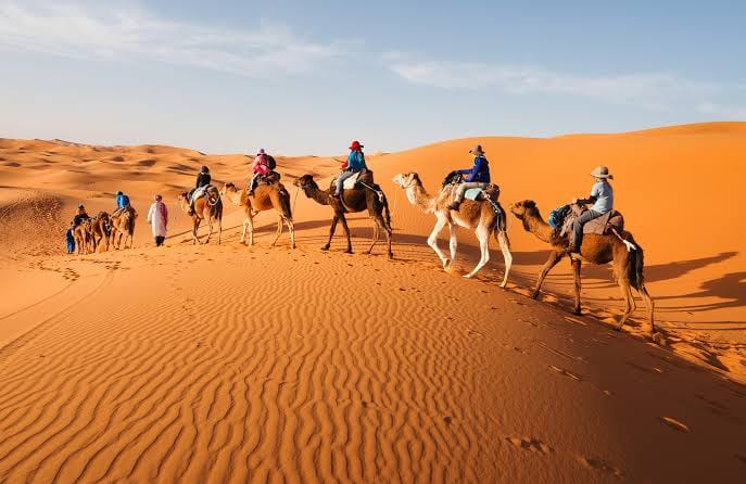 A line of riders on camels crosses the orange Erg Chebbi dunes led by a guide on foot