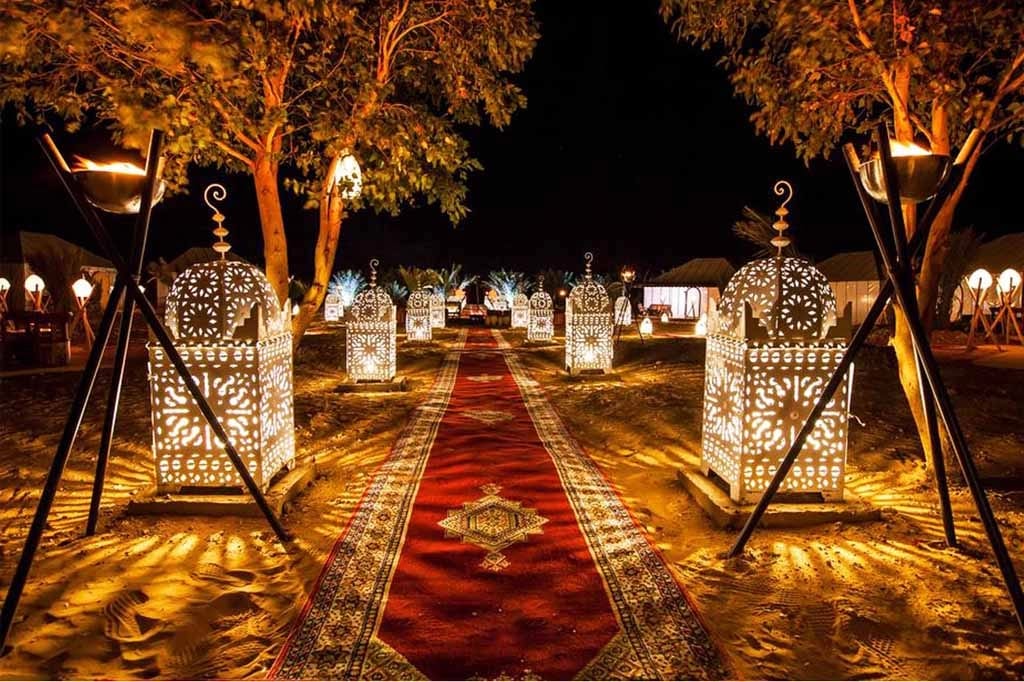Ornate white metal lanterns light a carpeted walkway through a Moroccan desert camp at night