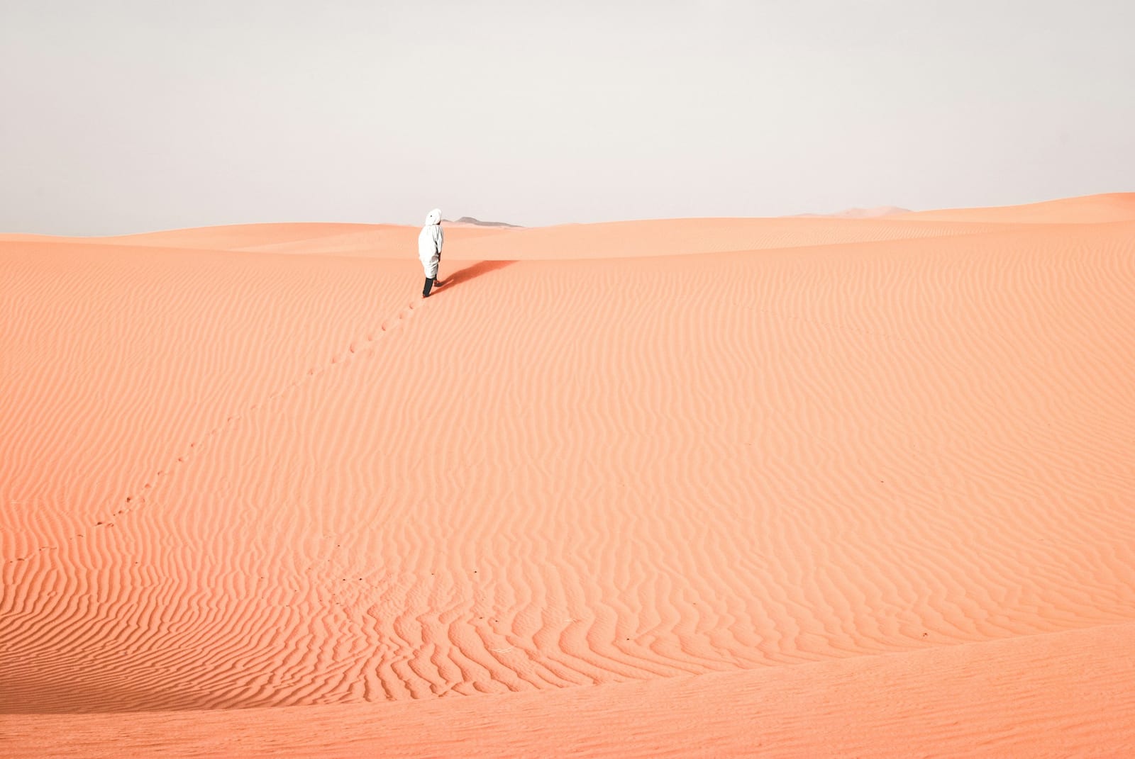 A lone figure in a white robe walks a dune ridge across empty Sahara sand
