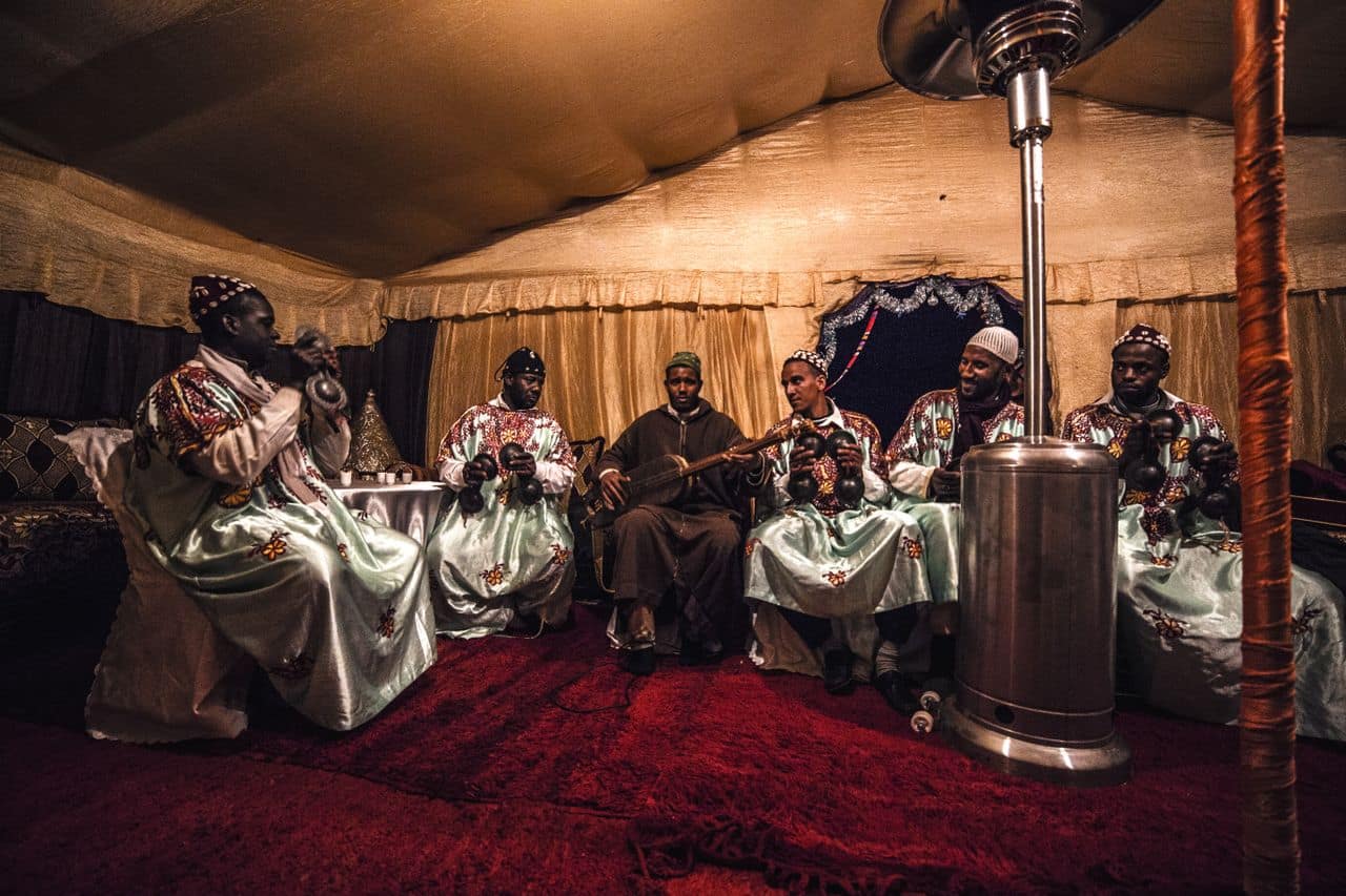 Gnawa musicians in embroidered robes play guembri and iron qraqeb inside a lamplit tent