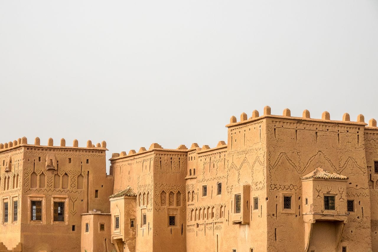Carved mud-brick kasbah towers with crenellated walls under a pale desert sky