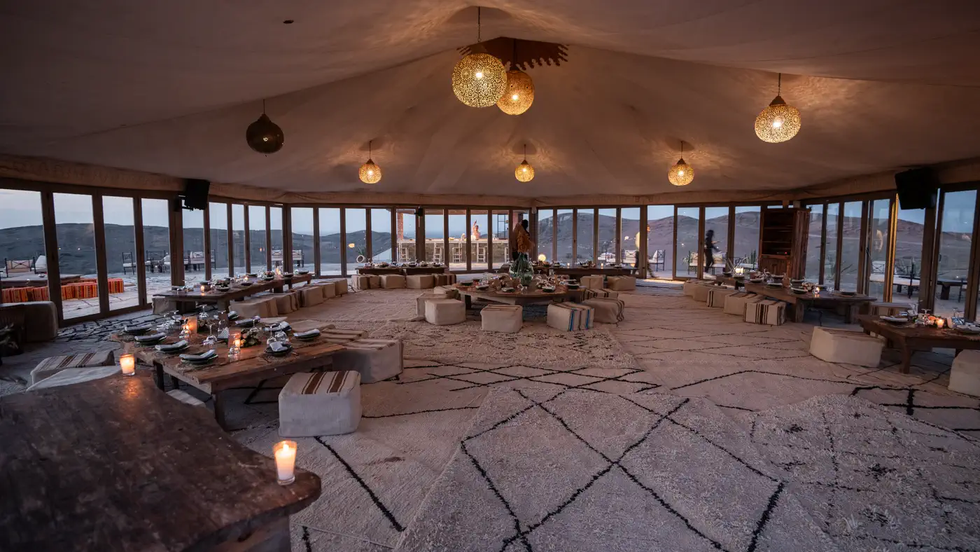 Glass-walled tent in the Agafay desert set with low tables and lanterns for a group dinner at dusk