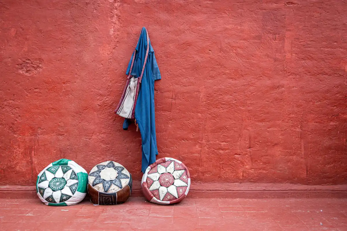 Handmade Moroccan leather poufs and a djellaba against the red ochre walls of Marrakech &mdash; artisan shopping and craft experiences sourced across our coverage area